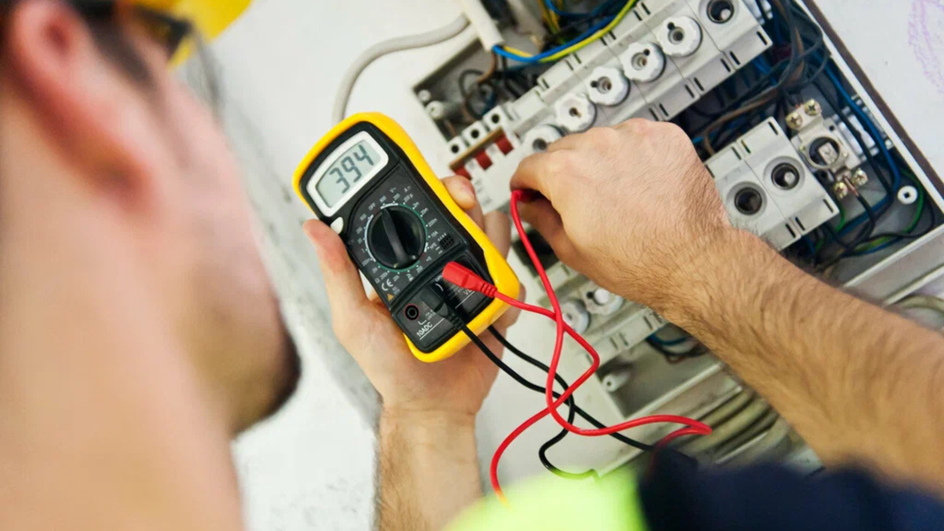 Electrician using a multimeter to test a circuit panel. Hands hold a yellow meter, wires connected.