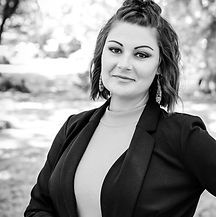 Woman in blazer and high-neck top poses outdoors with a slight smile, trees in the background.