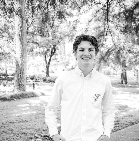 Smiling young man with braces outdoors, wearing a white button-down shirt, standing in front of trees.