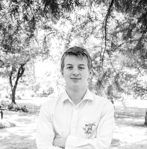 Young man with arms crossed, smiling in front of a tree with dappled sunlight.