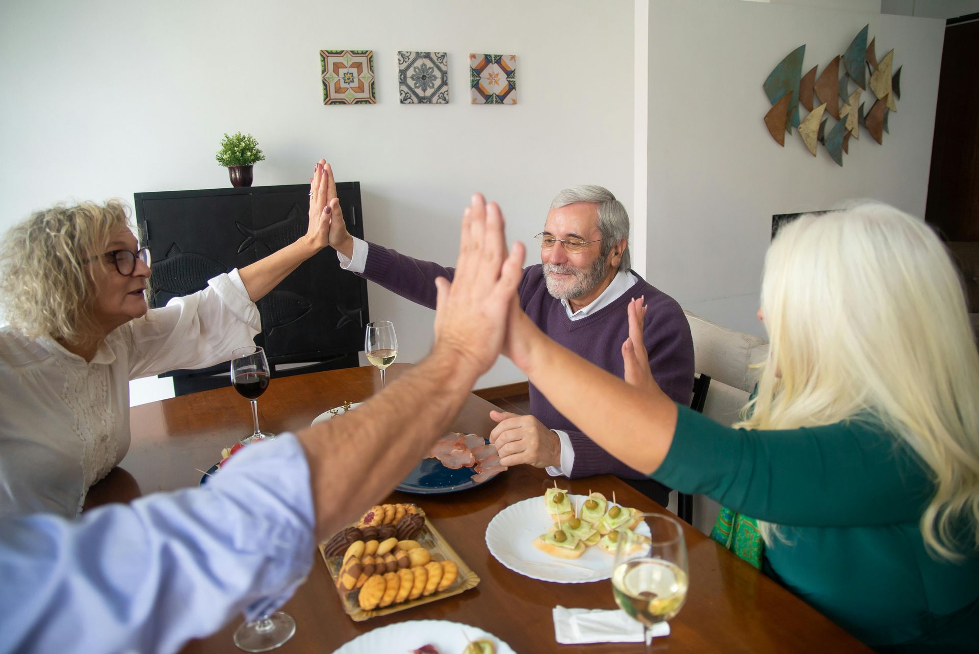 Four people at a table giving high fives. Older adults celebrating with food, drinks in a home setting.