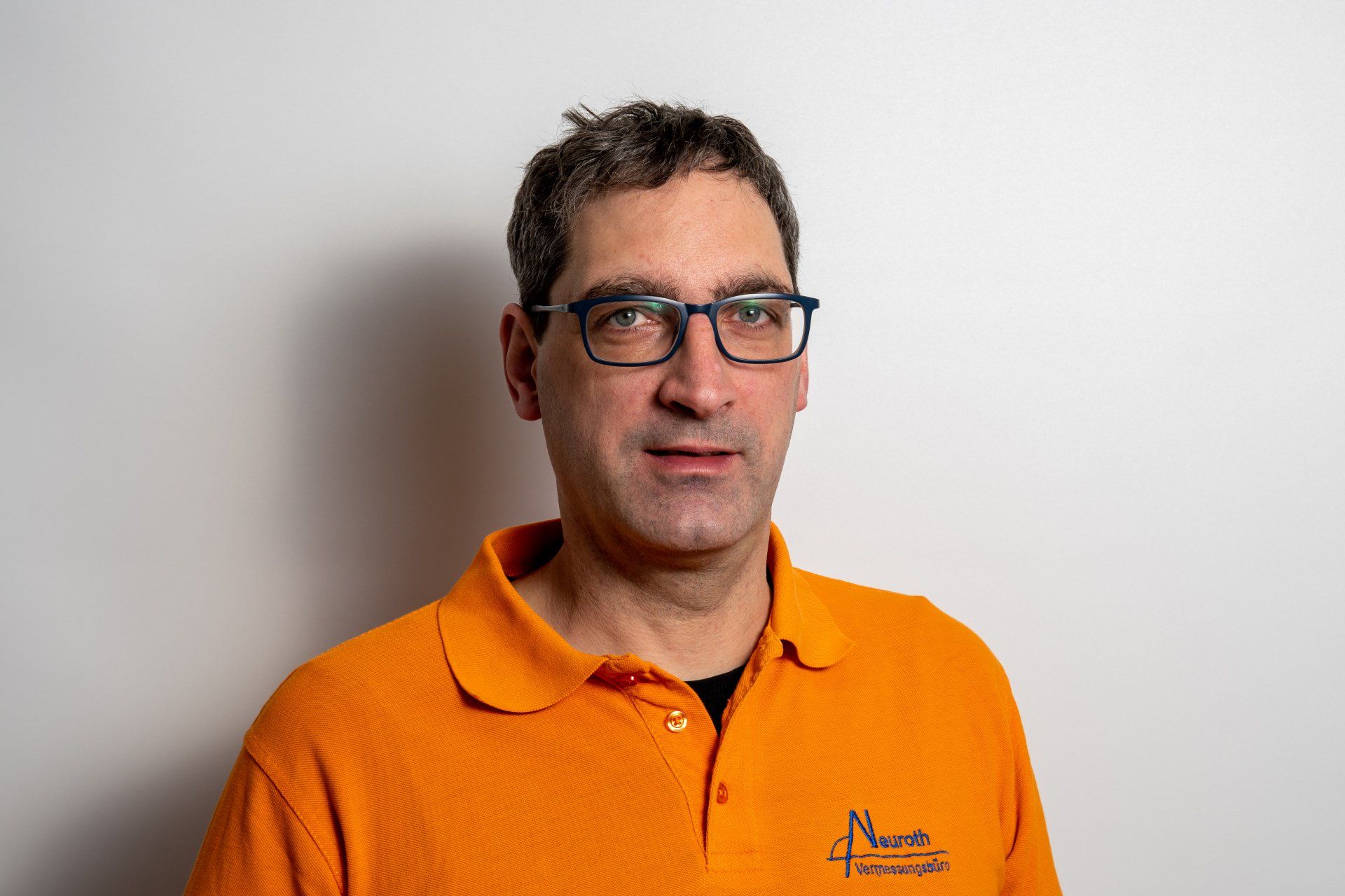 Ein Mann mit Brille und orangefarbenem Poloshirt steht vor einer weißen Wand.