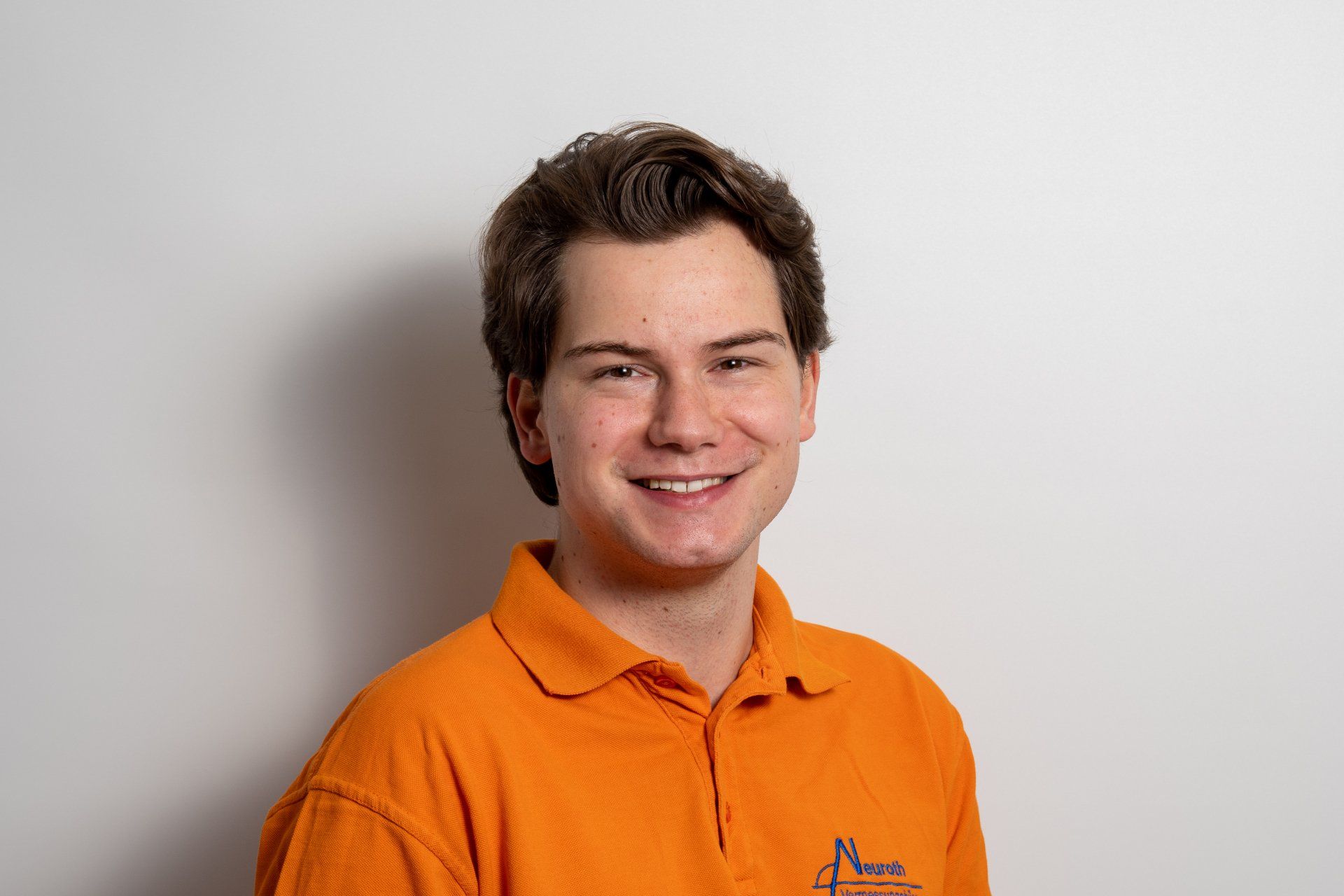 Ein junger Mann in einem orangefarbenen Poloshirt lächelt in die Kamera.