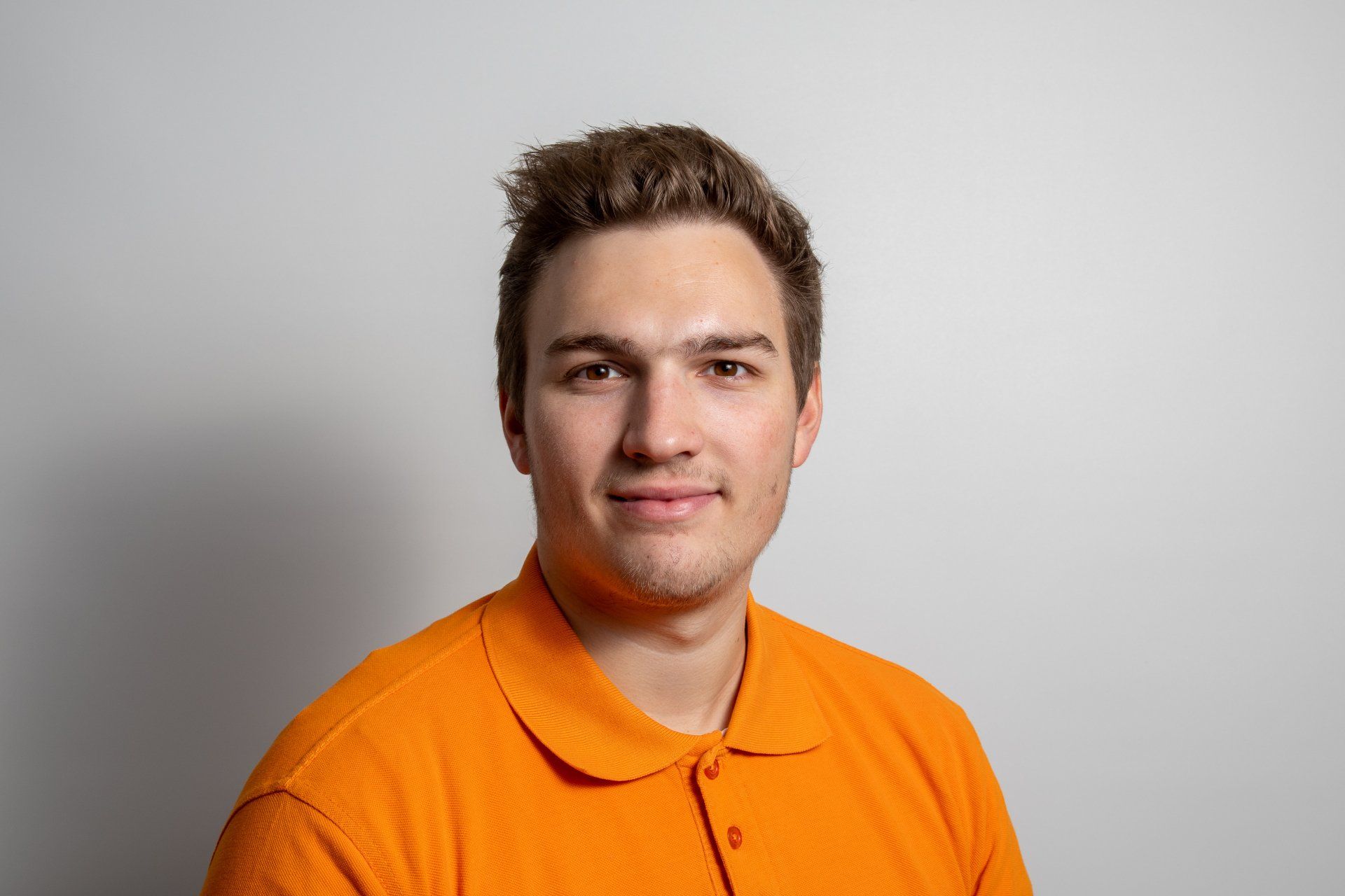 Ein junger Mann in einem orangefarbenen Poloshirt lächelt in die Kamera.