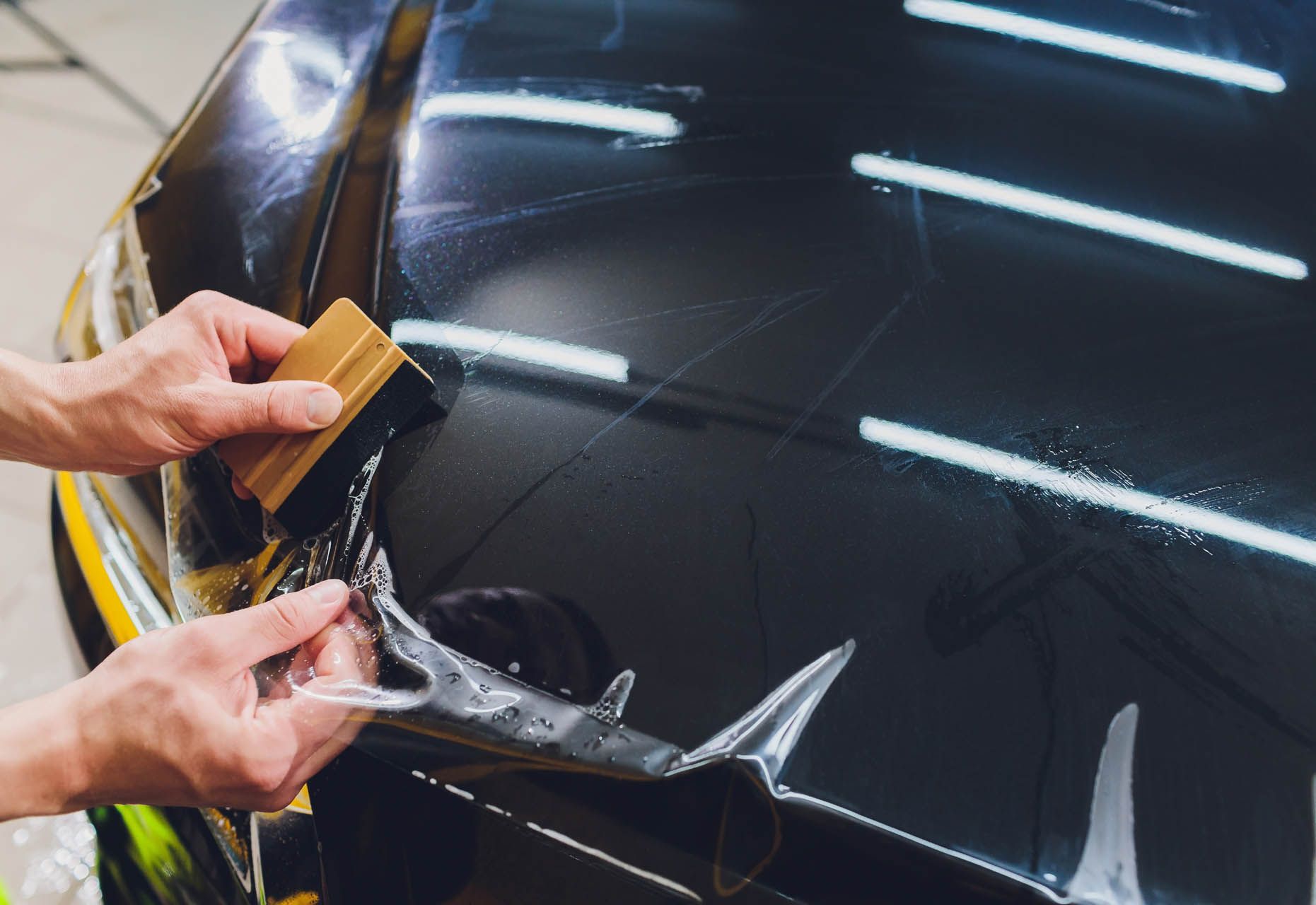 Hands applying protective film to a black car hood using a squeegee.