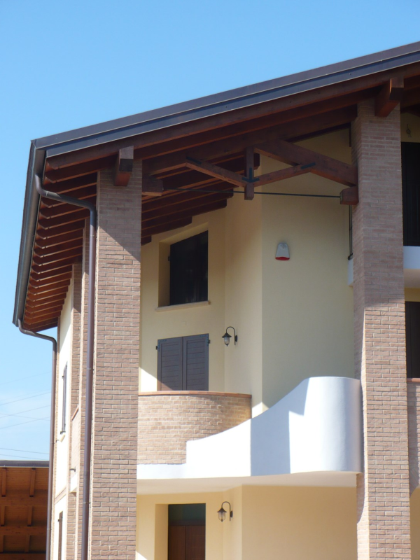 Edificio beige a due piani con colonne in mattoni, finiture in legno marrone, balcone e cielo azzurro terso.