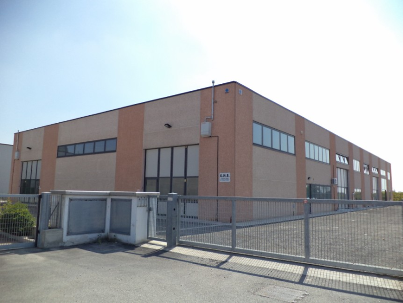 Un edificio industriale color marrone chiaro e rosso dietro un cancello di metallo grigio sotto un cielo limpido.