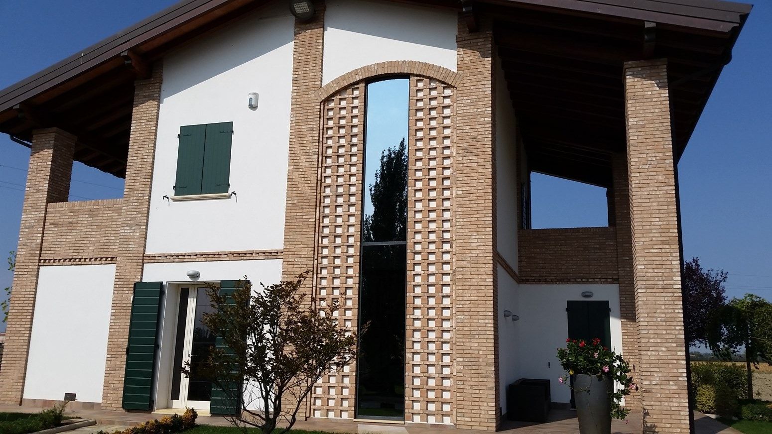 Casa a due piani in mattoni bianchi, con finestre alte e riflettenti e persiane verdi, sotto un cielo azzurro.