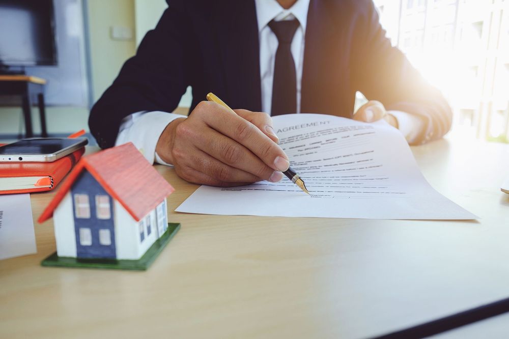 Persona in giacca e cravatta che firma documenti immobiliari, con un modellino di casa sulla scrivania.
