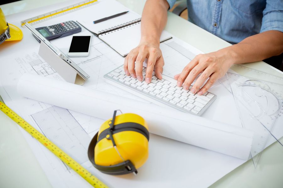 Persona che digita su una tastiera seduta su una scrivania ricoperta di progetti, attrezzi e una calcolatrice.