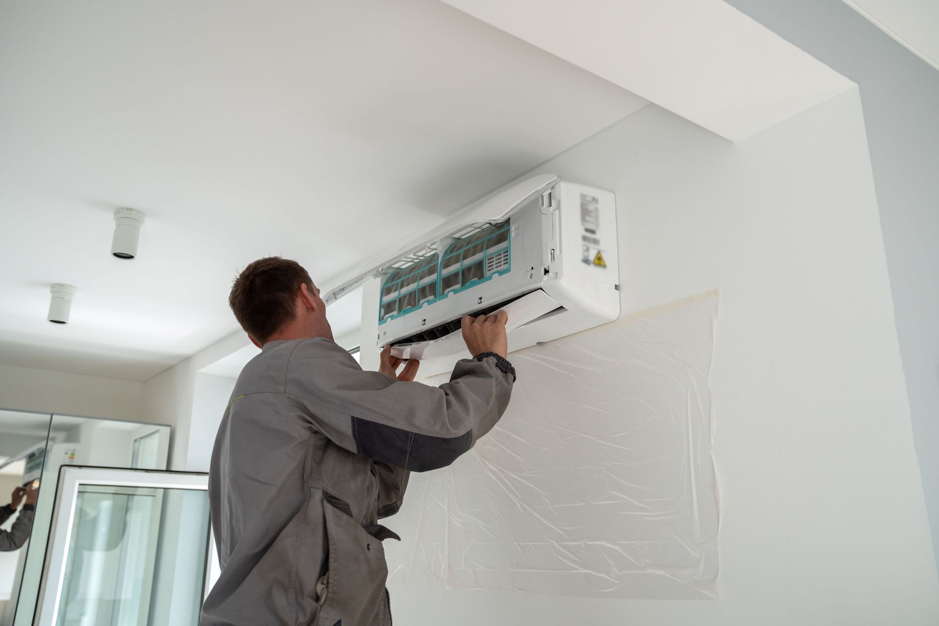 Man in grey uniform installing a white air conditioning unit on a white wall indoors.