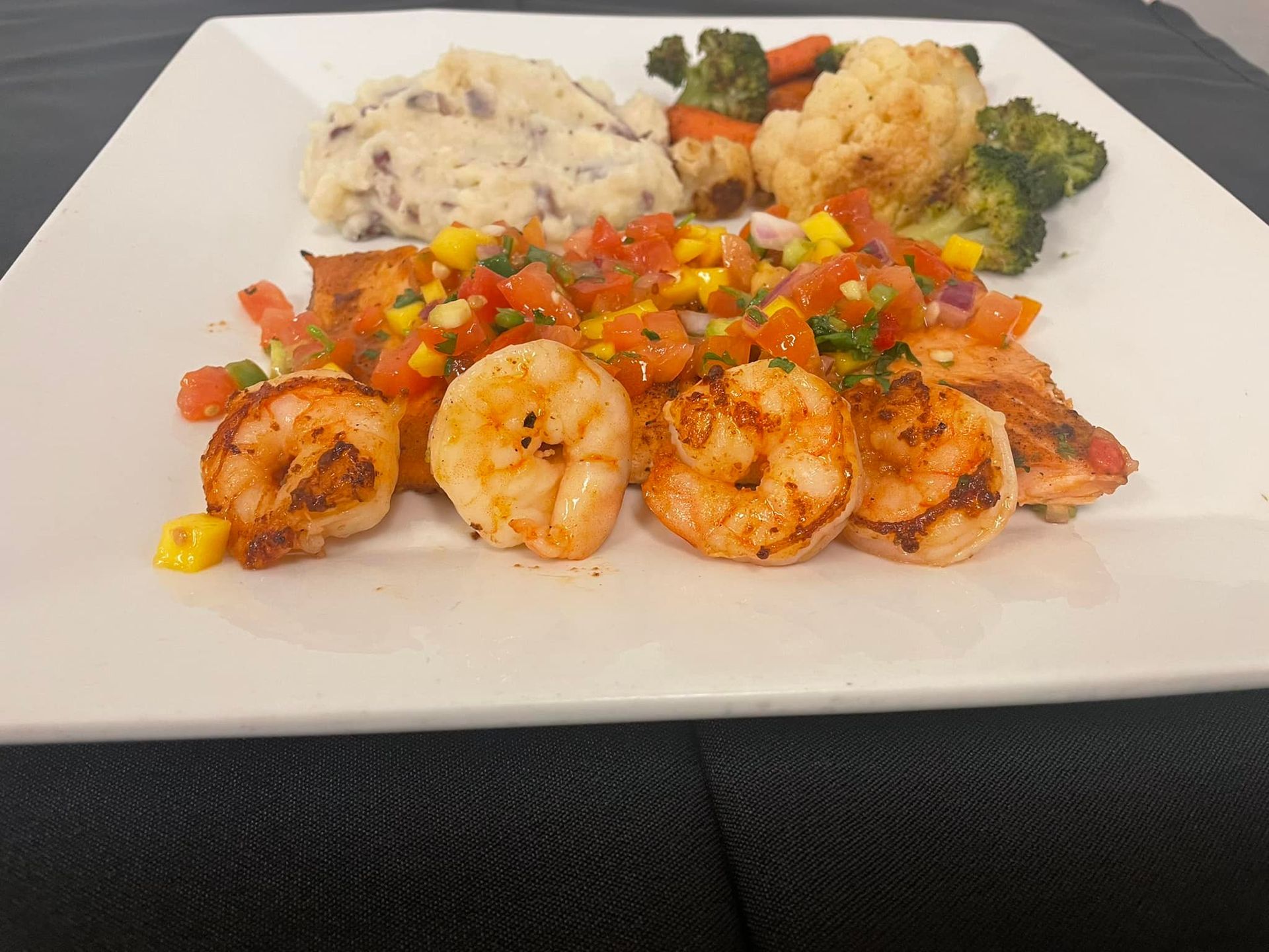 A white plate holding grilled shrimp topped with salsa, a side of mashed potatoes, and roasted cauliflower and broccoli.