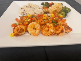 A white plate holding grilled shrimp topped with salsa, a side of mashed potatoes, and roasted cauliflower and broccoli.