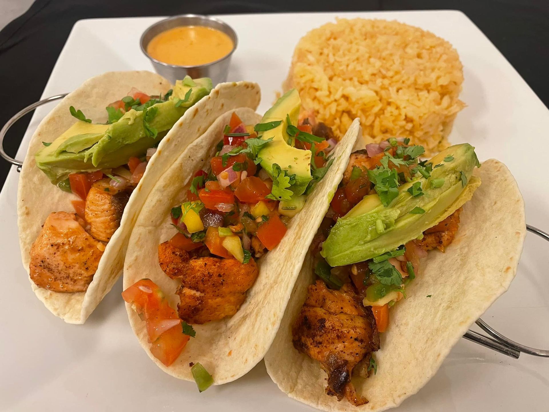 Three fish tacos topped with avocado and salsa, served with a side of yellow rice and a cup of sauce on a white plate.