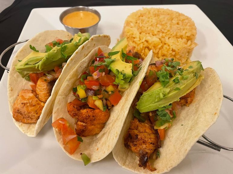 Three fish tacos topped with avocado and salsa, served with a side of yellow rice and a cup of sauce on a white plate.
