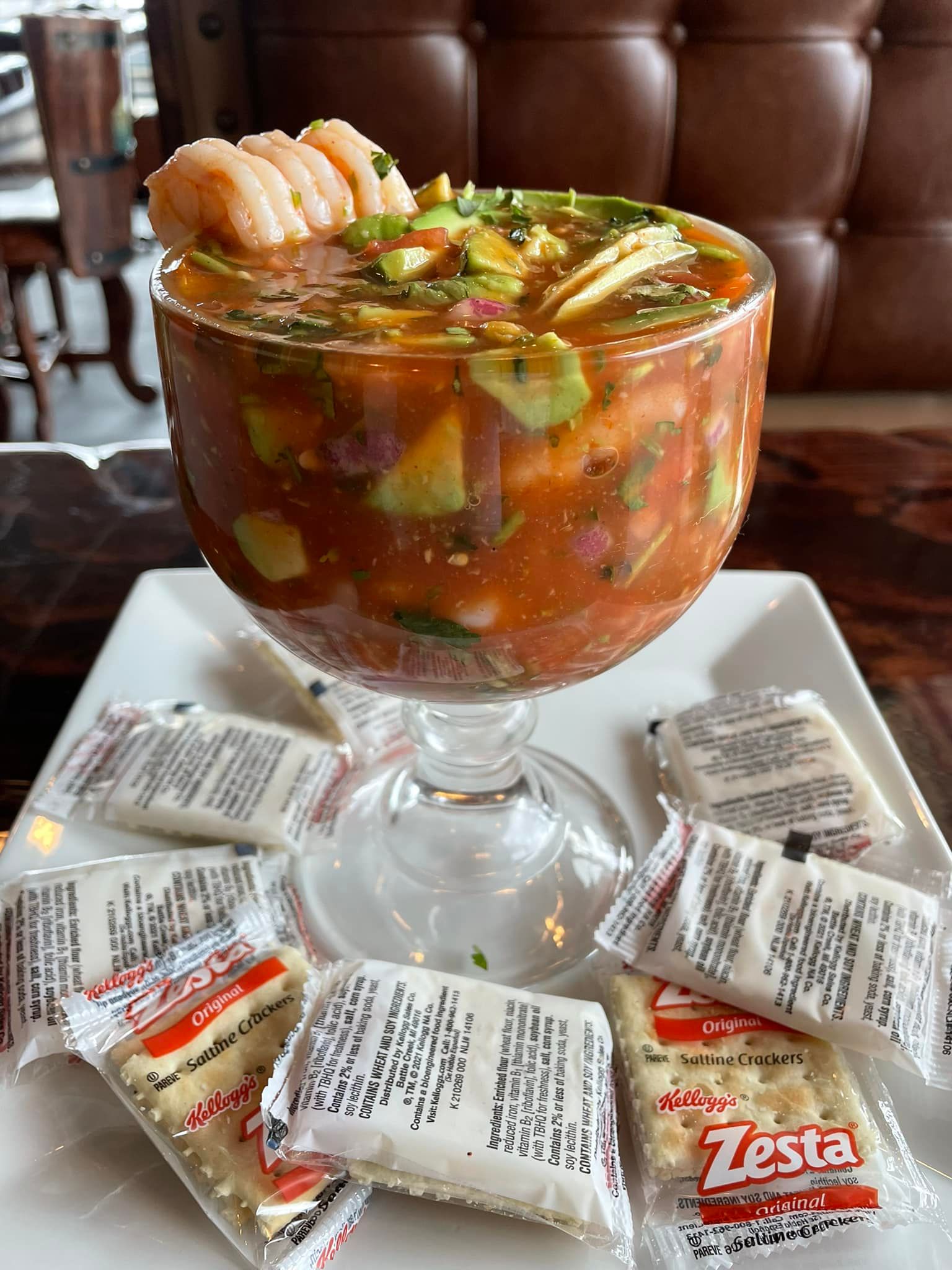 A shrimp cocktail served in a large glass goblet on a white plate surrounded by several individual Zesta cracker packs.