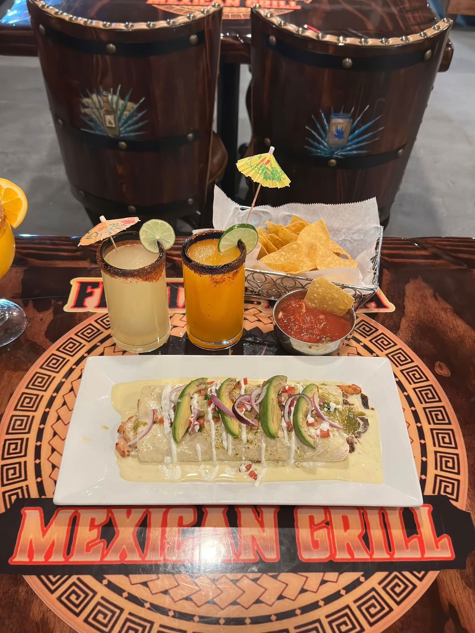 A burrito topped with avocado sits on a restaurant table with two colorful drinks, chips, and salsa.