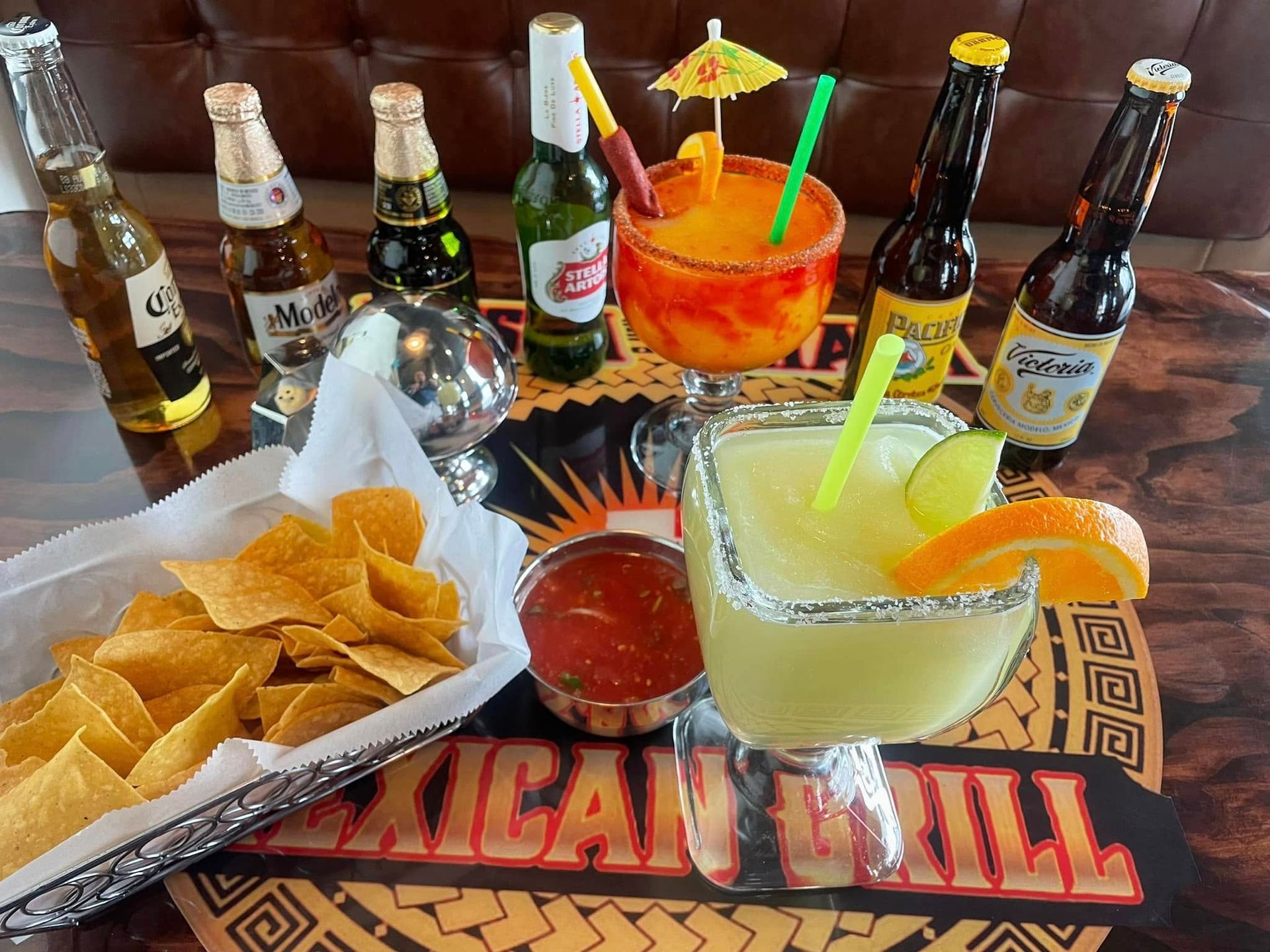 A basket of tortilla chips, salsa, two colorful margaritas, and several beer bottles on a Mexican Grill restaurant table.
