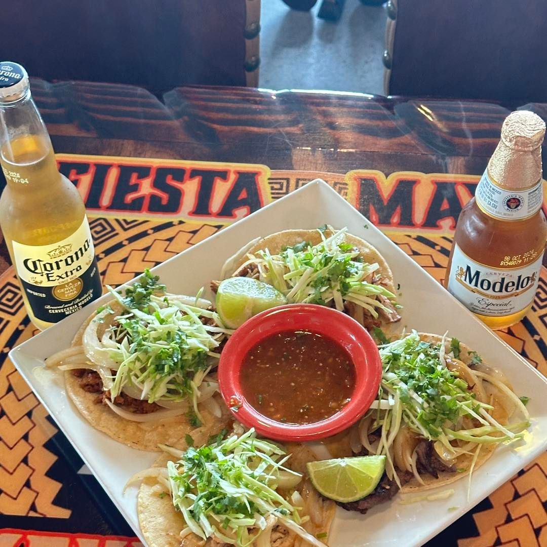 A white square plate with four tacos topped with shredded cabbage and cilantro, a red bowl of salsa, and two beer bottles.