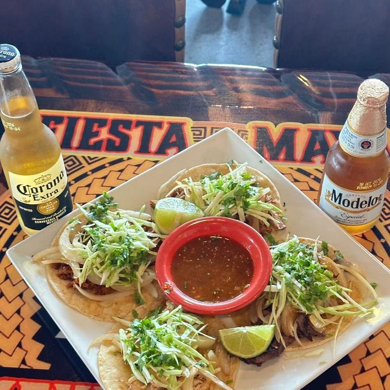 A white square plate with four tacos topped with shredded cabbage and cilantro, a red bowl of salsa, and two beer bottles.