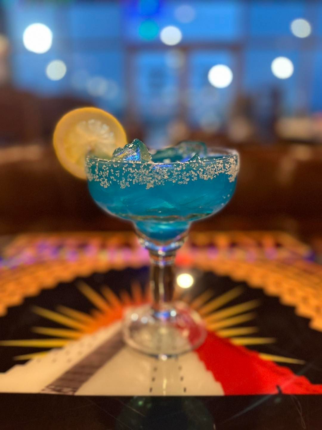 A blue margarita in a salt-rimmed glass with a lemon slice garnish, sitting on a colorful, patterned table.