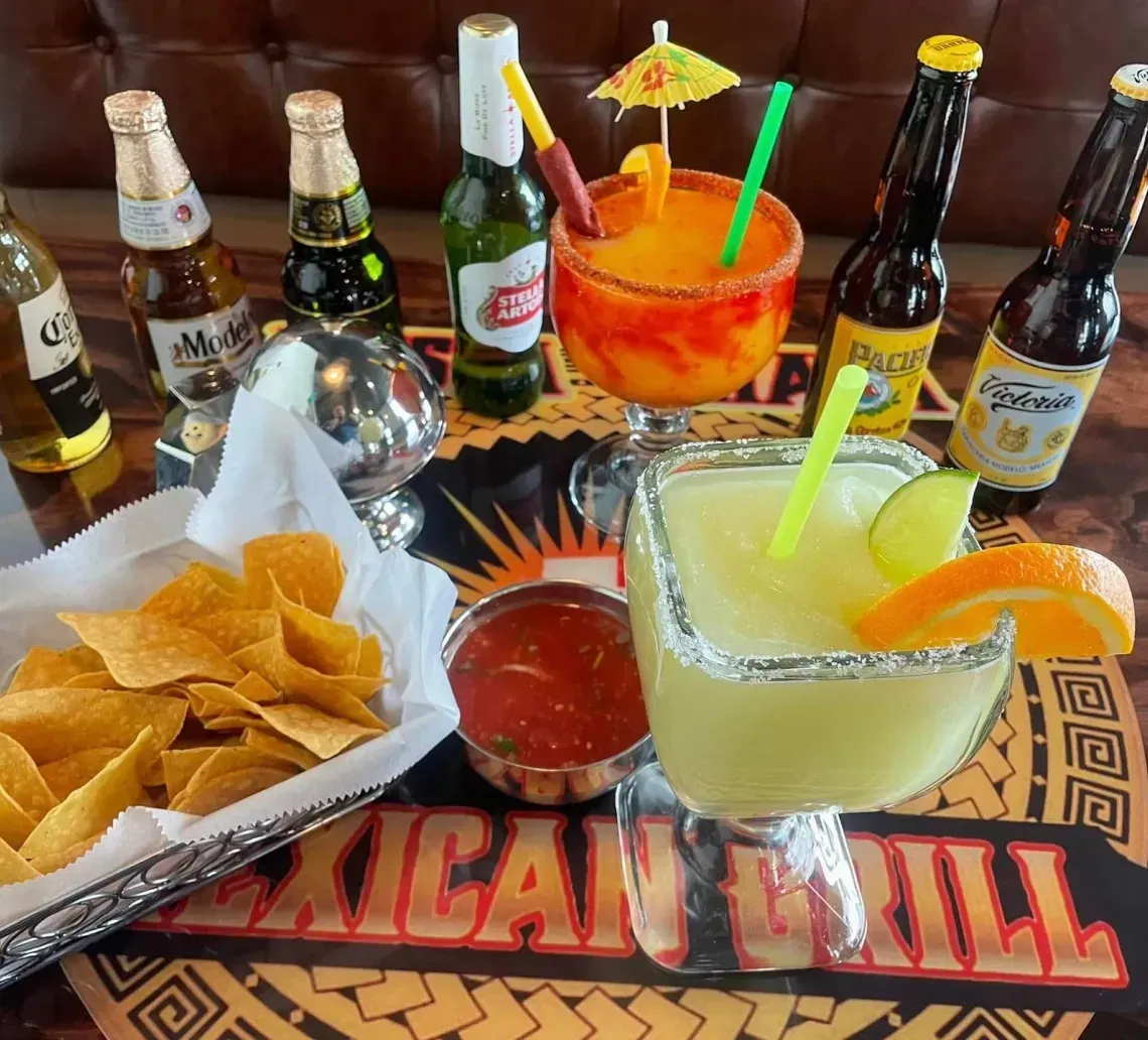 A basket of tortilla chips, salsa, two colorful margaritas, and several beer bottles on a Mexican Grill restaurant table.