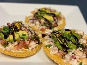 Three shrimp ceviche tostadas topped with avocado slices, fresh herbs, and a balsamic glaze on a white plate.