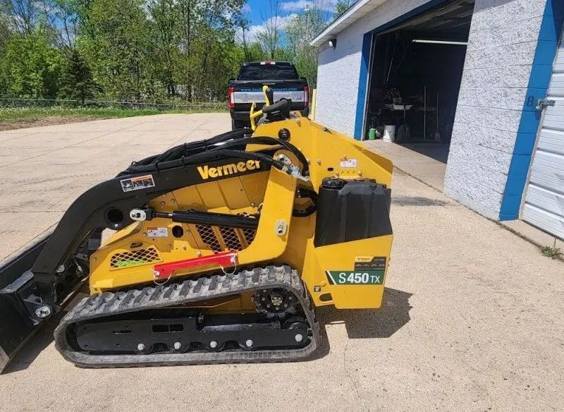 Yellow and black Vermeer mini skid steer parked outside an open garage with a truck nearby