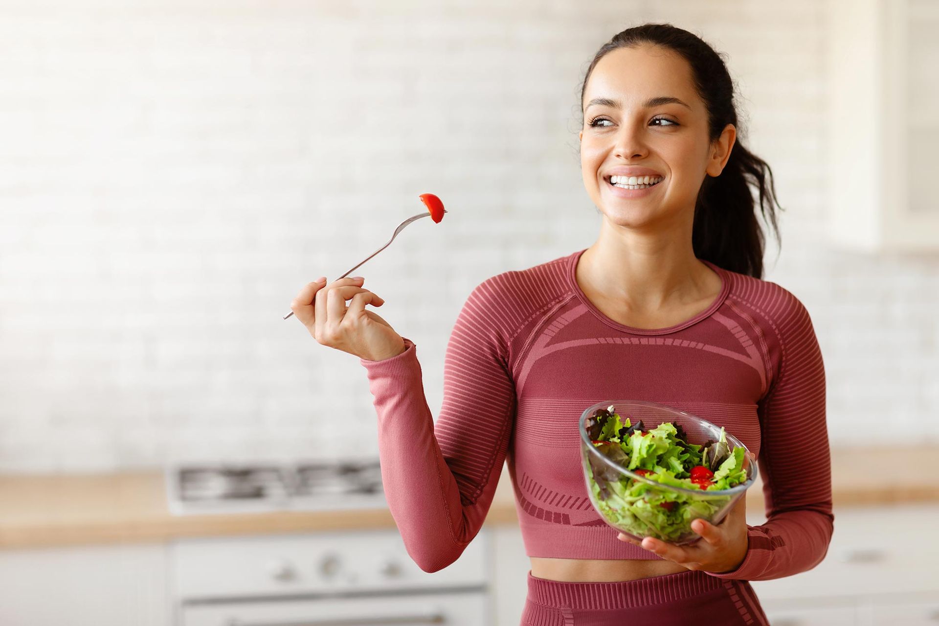 Woman in workout clothes smiling, eating salad in a kitchen.