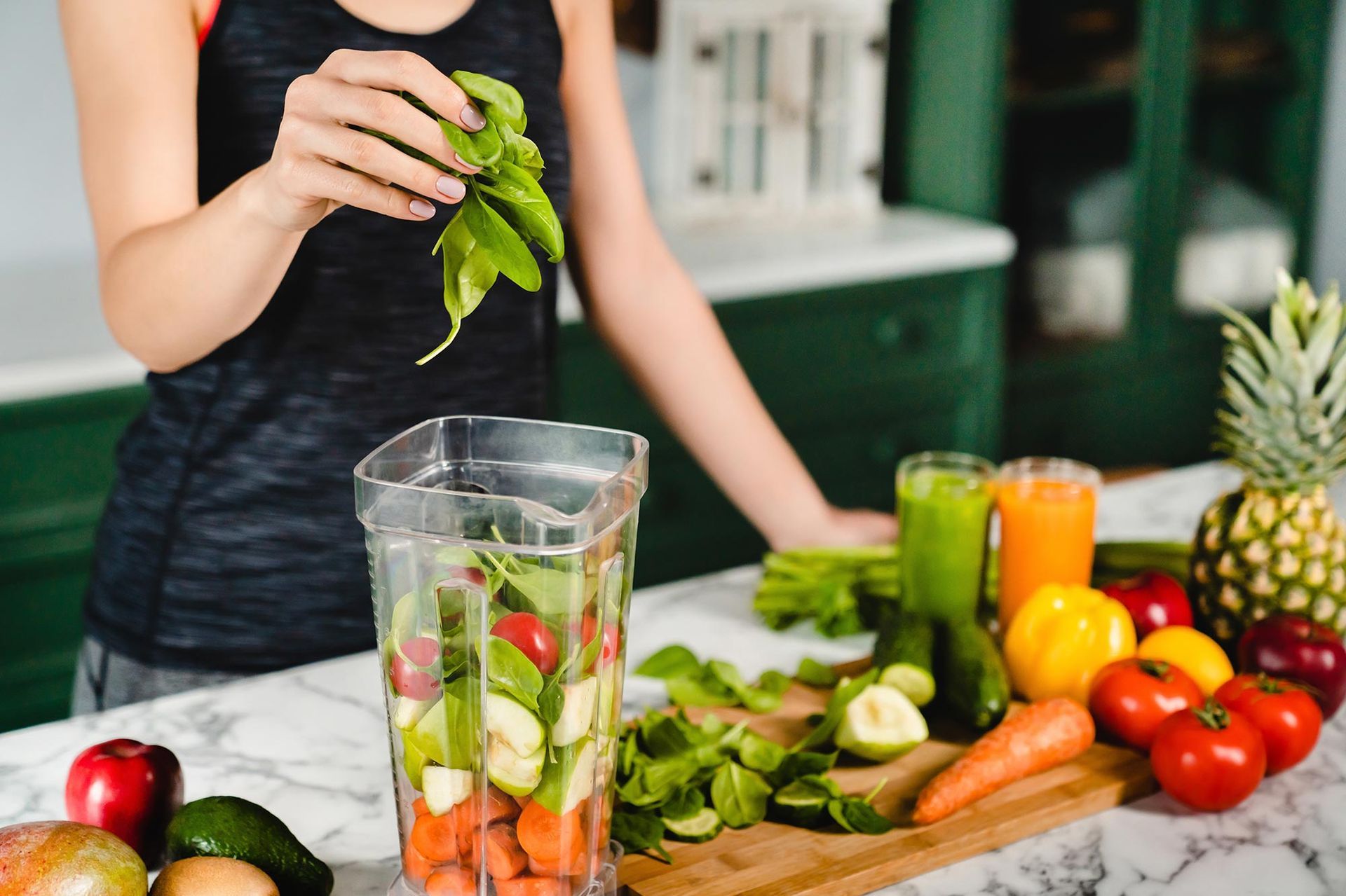 Woman adding spinach to a blender filled with fruits and vegetables for a smoothie, with more produce nearby.