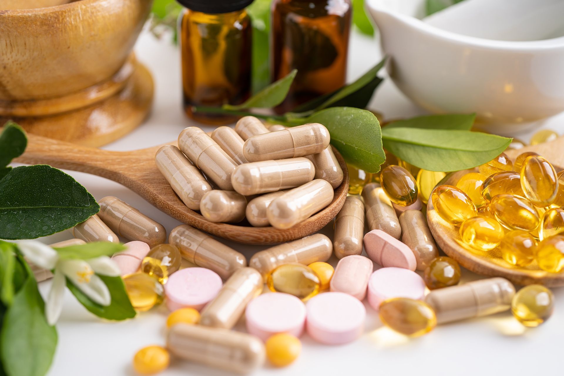 Capsules, pills, and supplements on wooden spoons, with essential oil bottles and foliage.