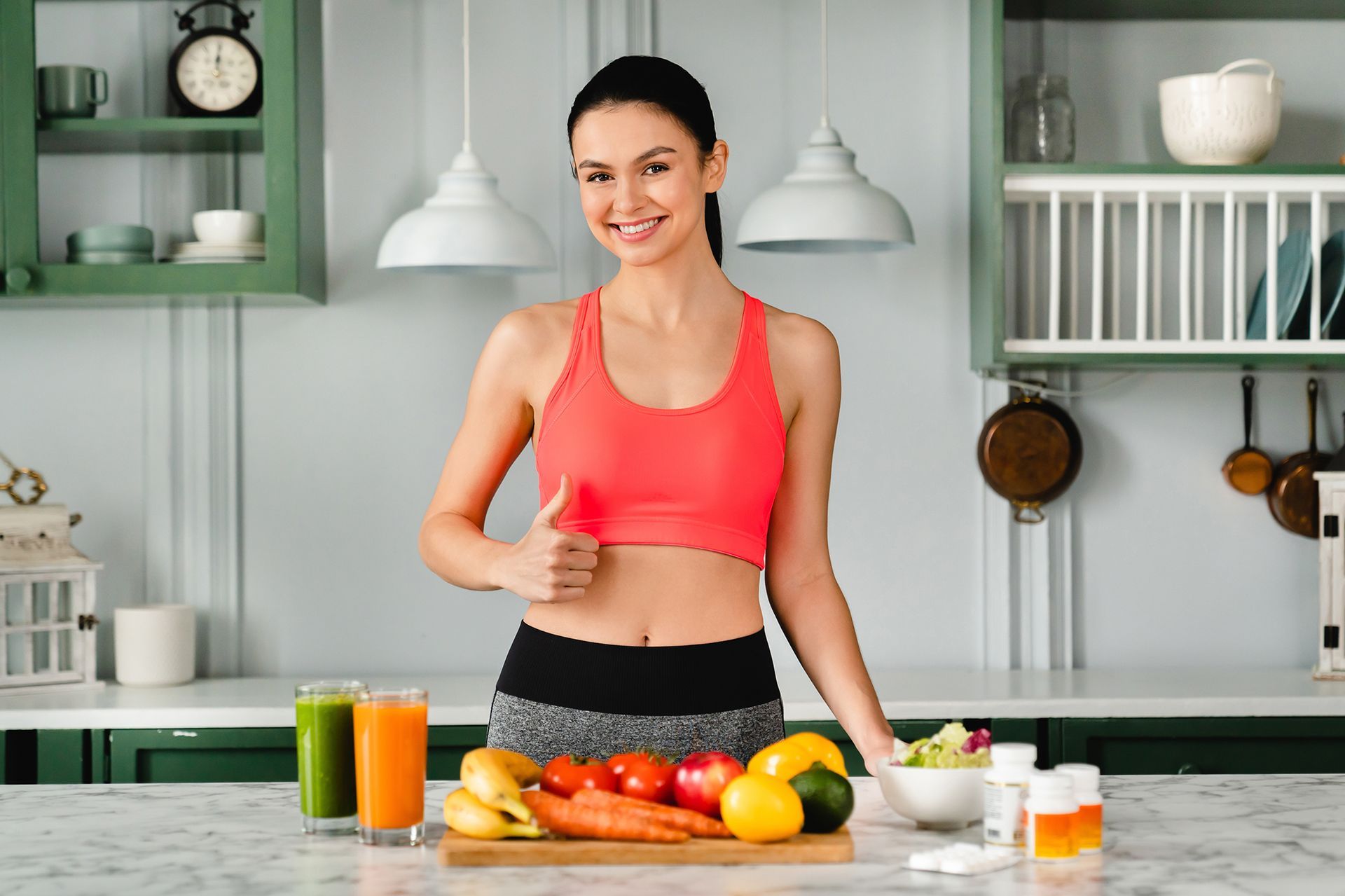 Woman in workout clothes gives thumbs up in a kitchen with fruits, vegetables, and smoothies.