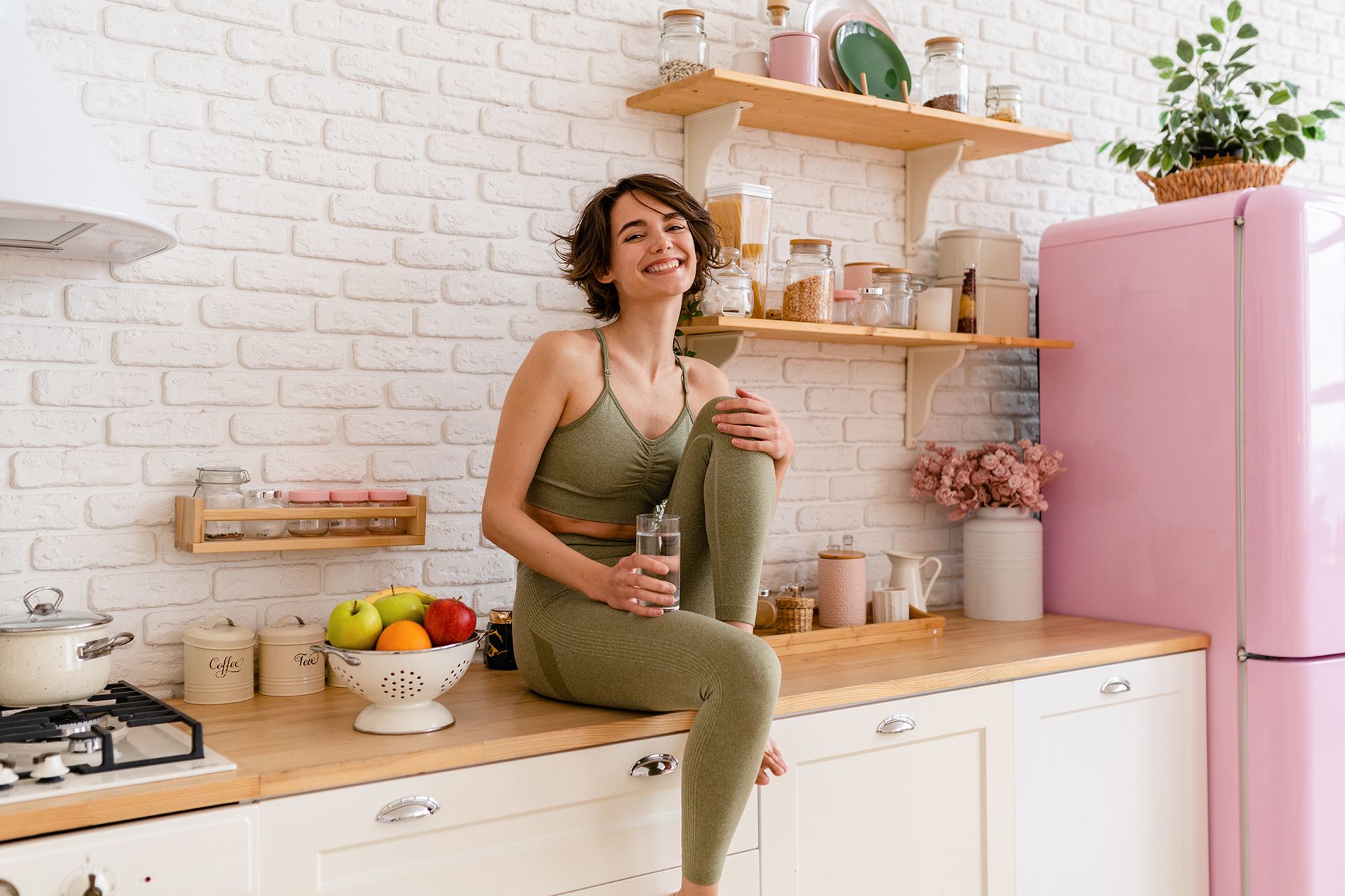 Woman in olive green workout clothes smiles in a kitchen, holding a glass of water.