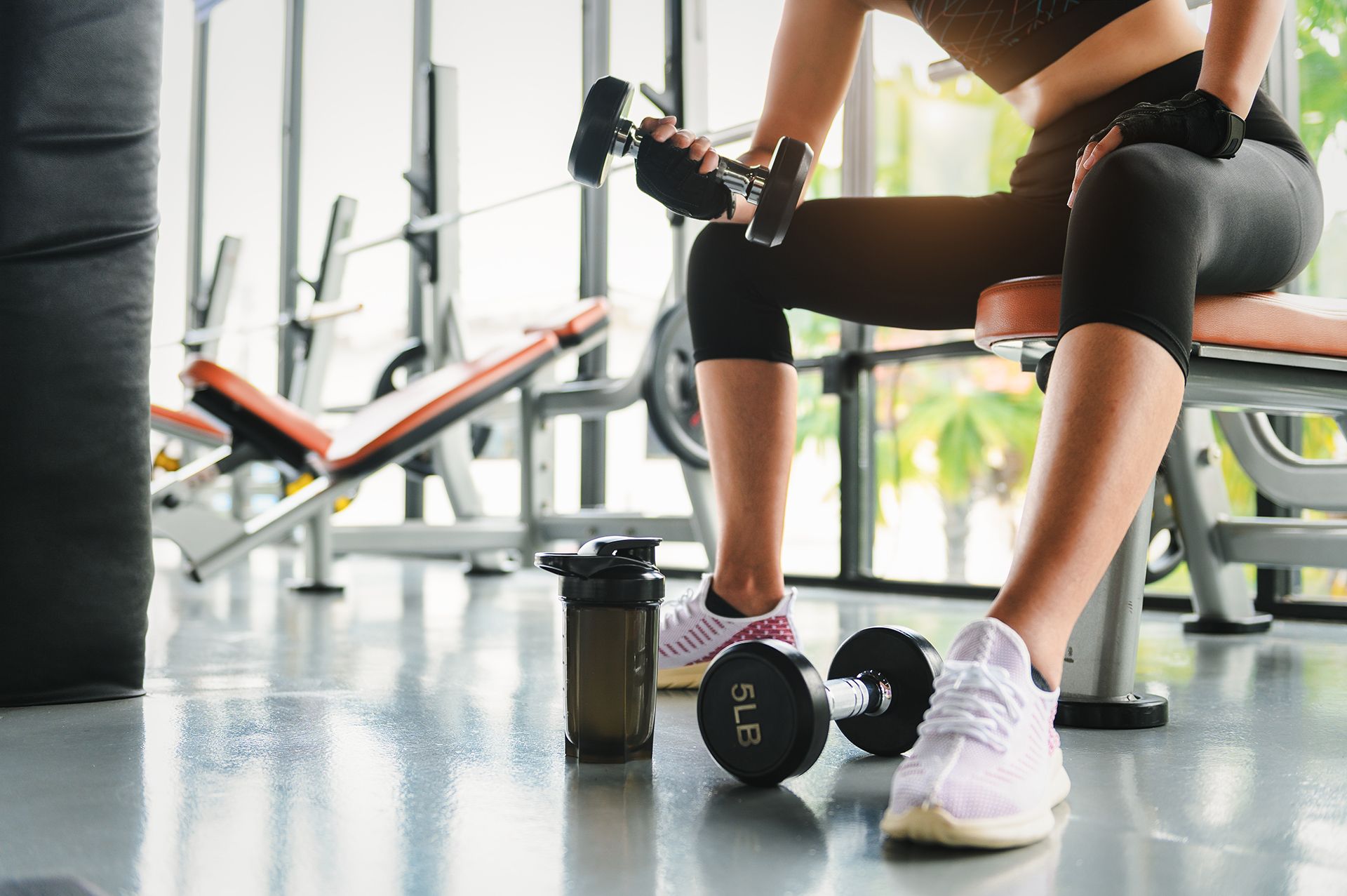Woman in workout clothes lifting dumbbells on a bench in a gym.