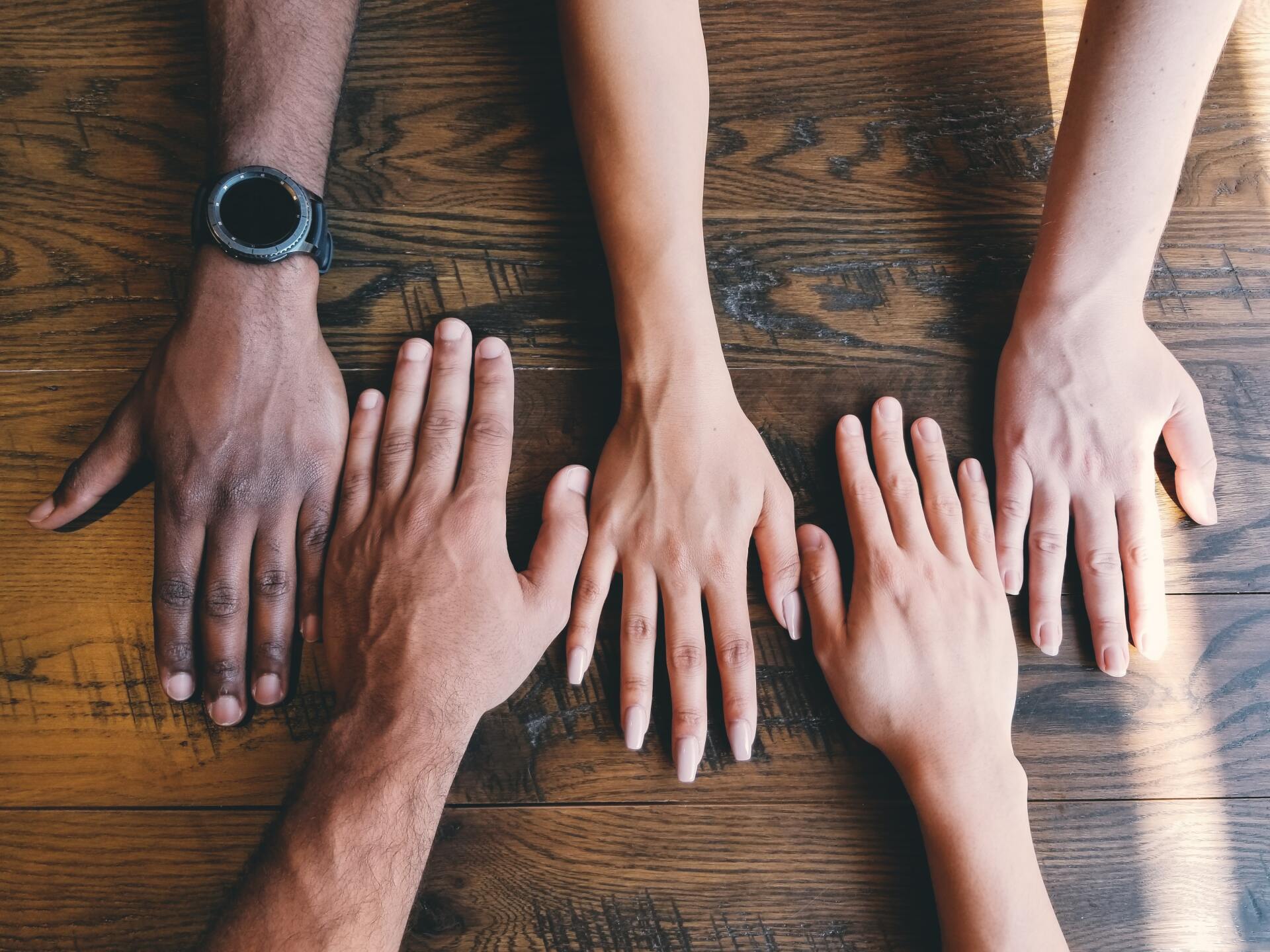 five hands of different ethnicities on a wooden table one wearing a black watch