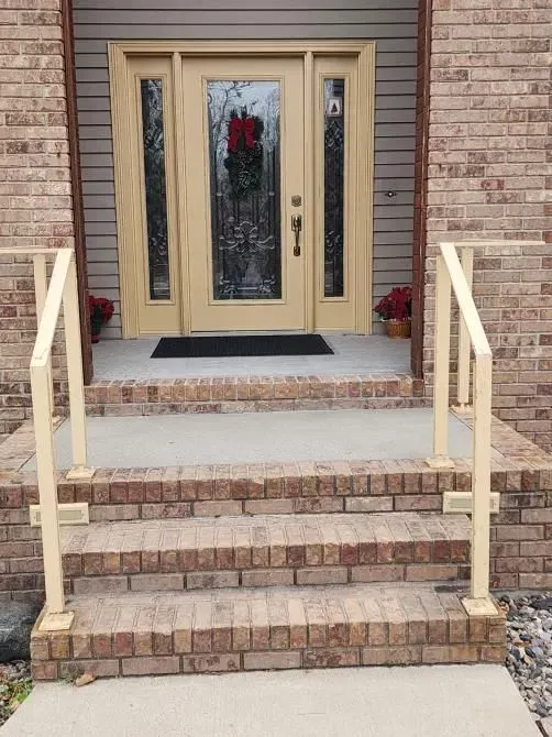 Beige front door with sidelights, red brick steps, and beige handrails.