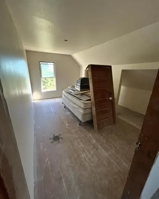 Attic room with mattress, stacked items, and a wooden door. Sloped ceiling, unfinished walls, and a window.