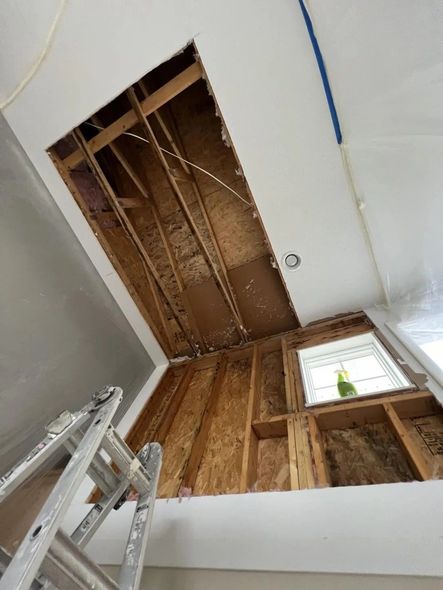 Interior view of ceiling with a large hole revealing wood frame construction; a ladder leans against the wall.