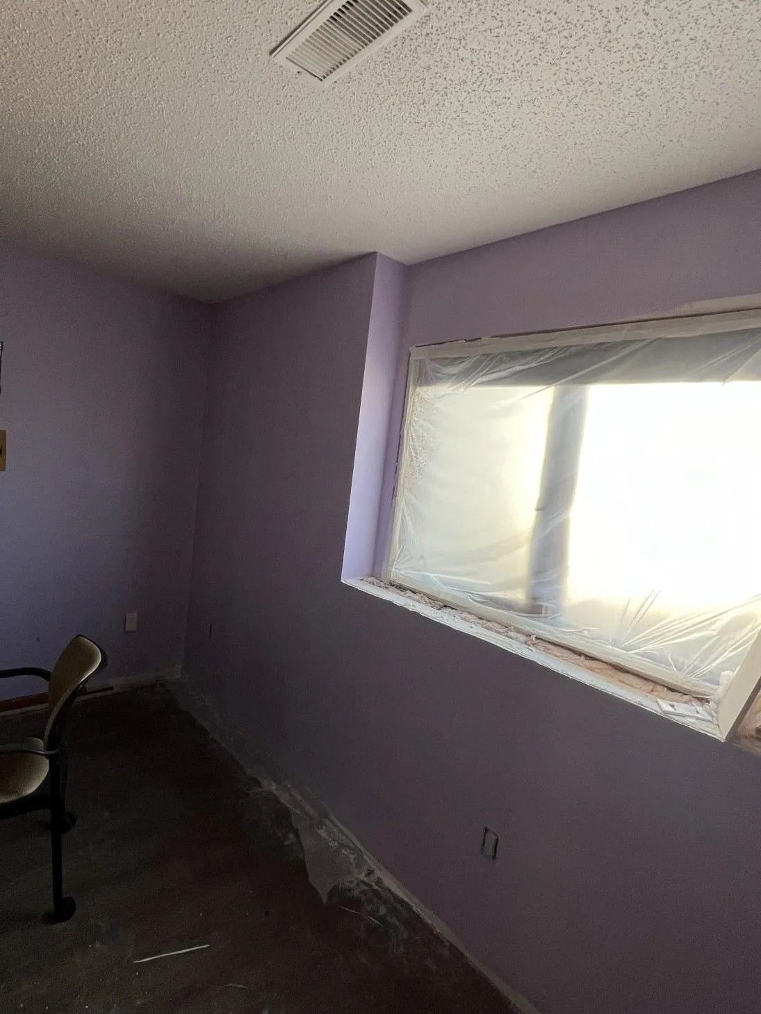 Purple-painted room with window covered in plastic, chair in the corner, and a textured ceiling.