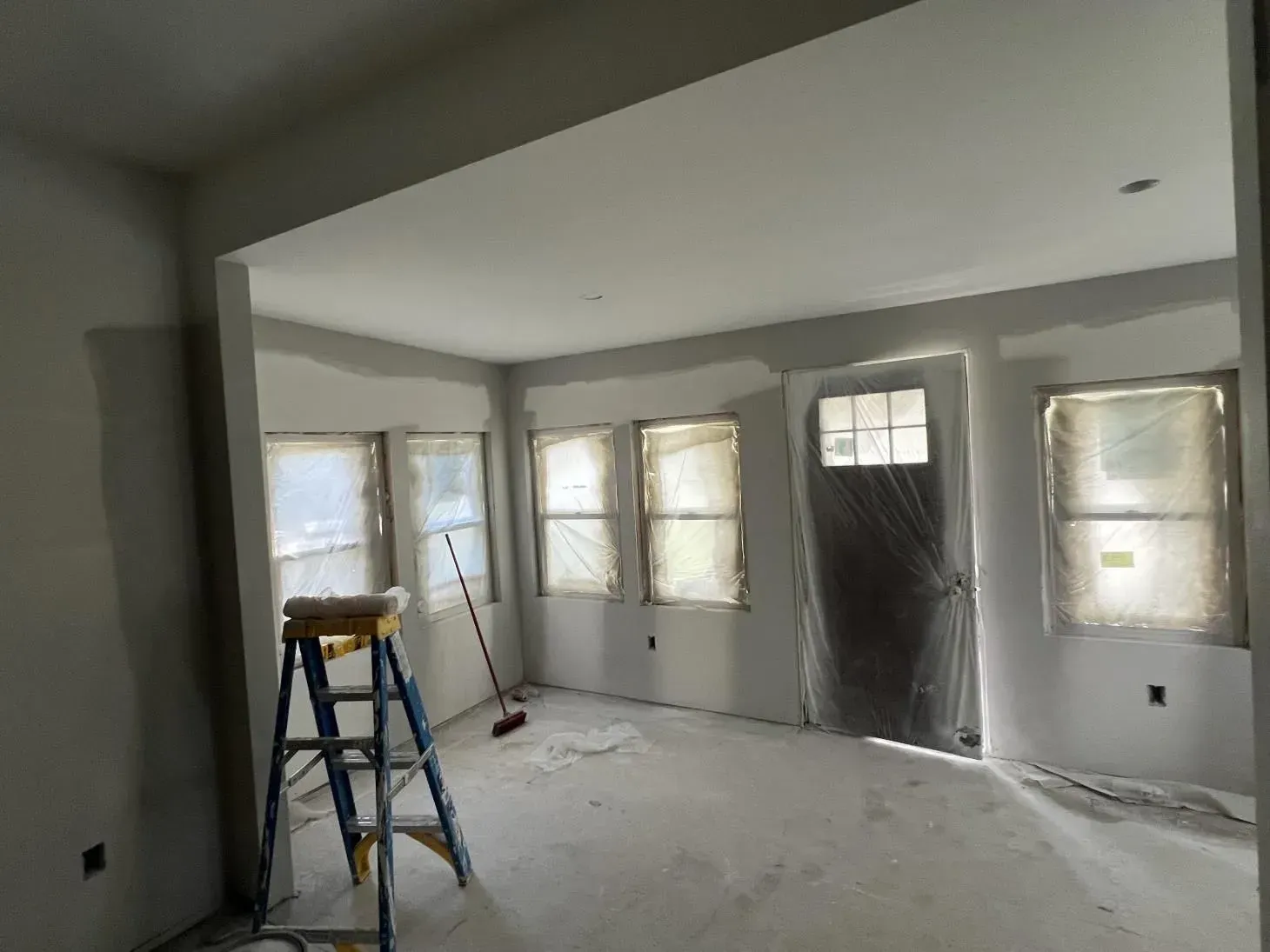 Interior room under construction, with windows and door covered in plastic, ladder, and drywall.