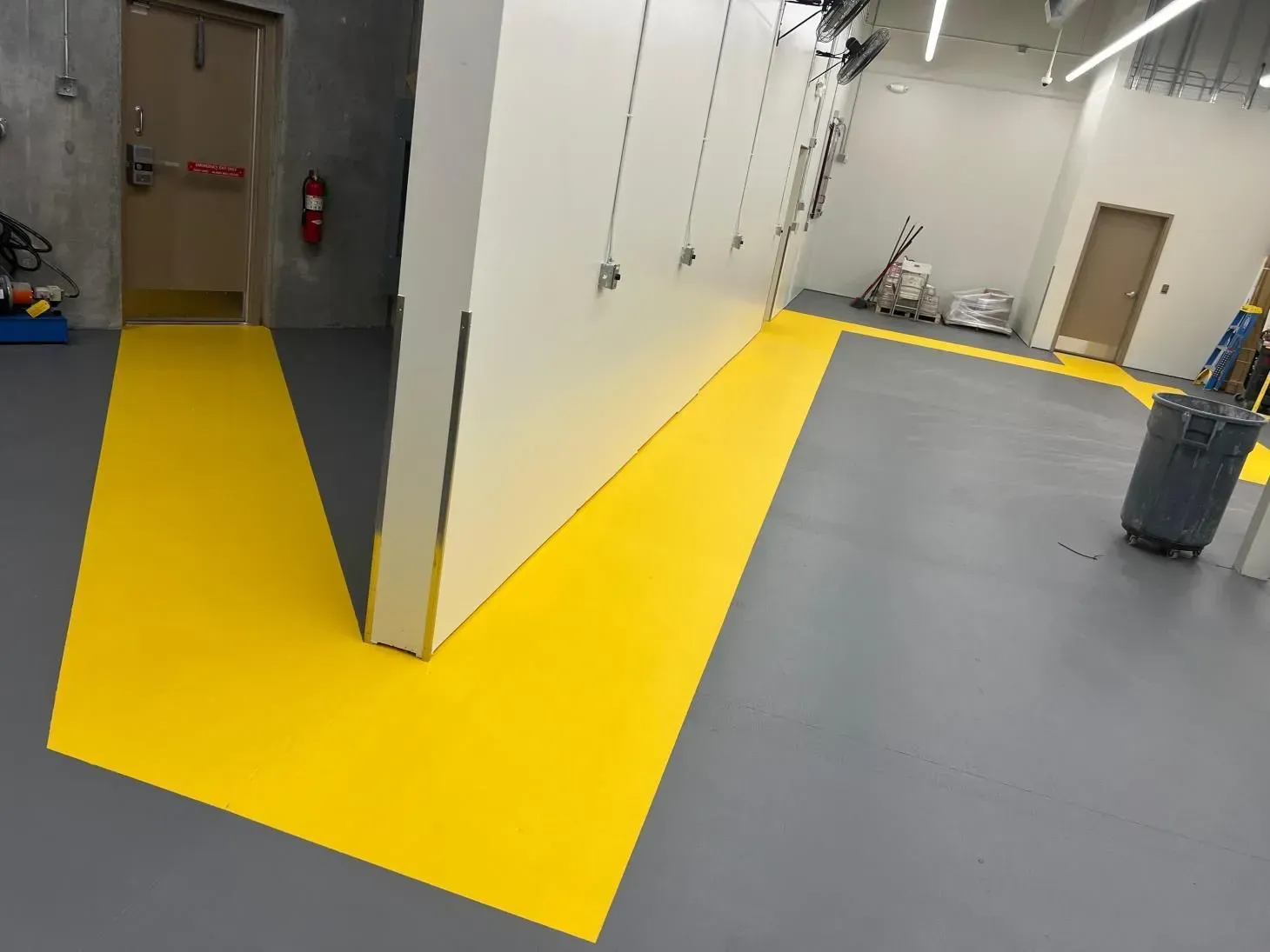 Yellow painted pathway on gray floor leads to a door, running alongside a white wall in a room.