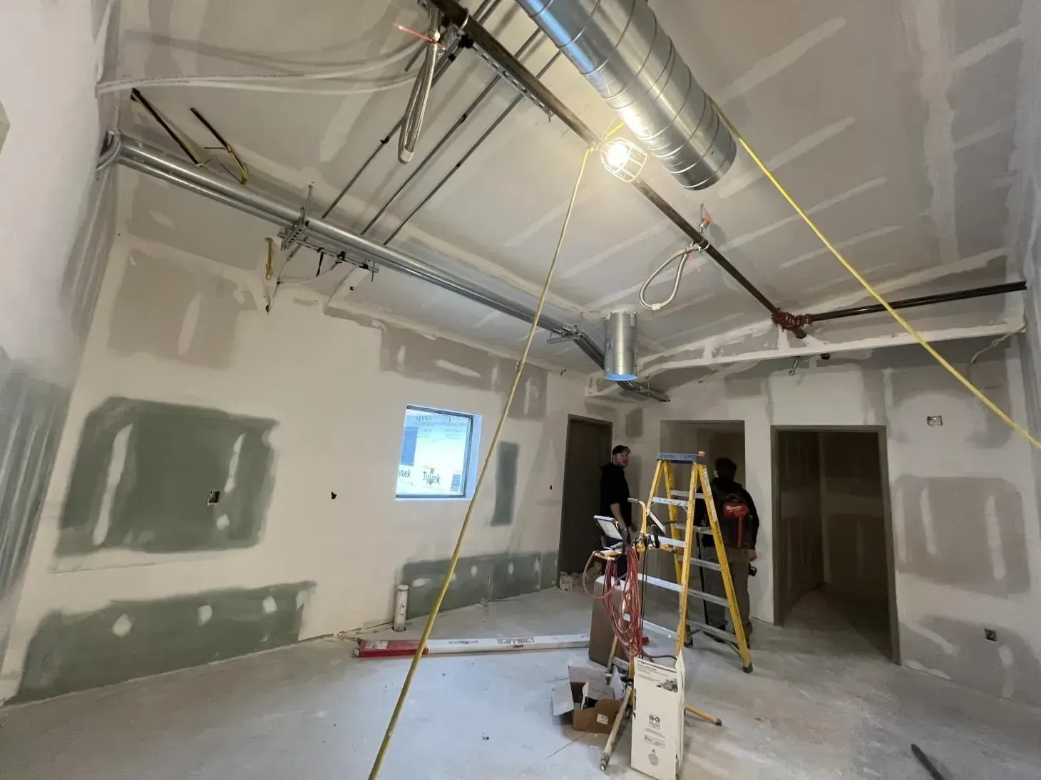 Interior construction site: drywall on walls and ceiling, two people, ladder, ductwork.