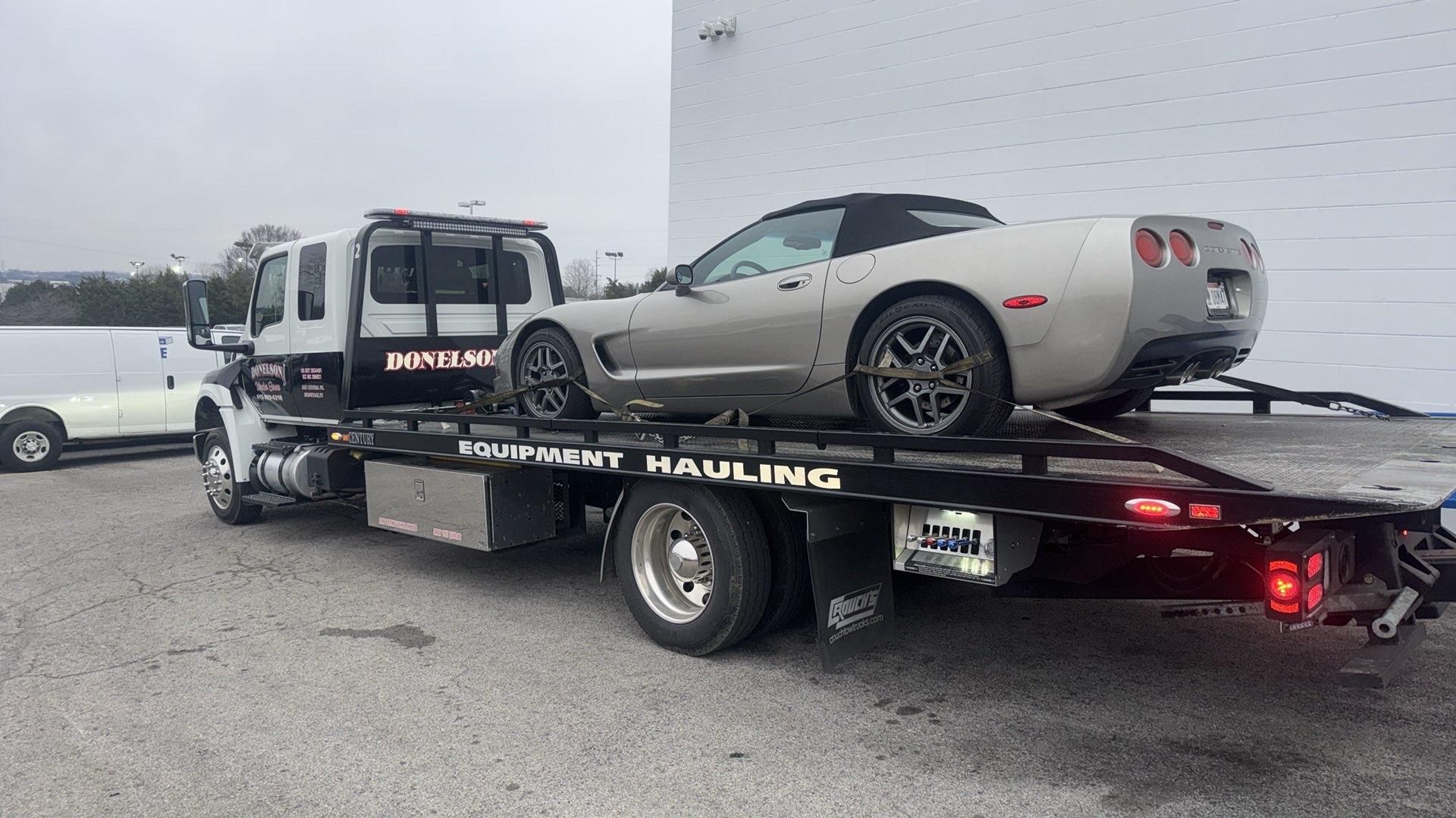 Silver convertible Corvette on a flatbed tow truck for equipment hauling.