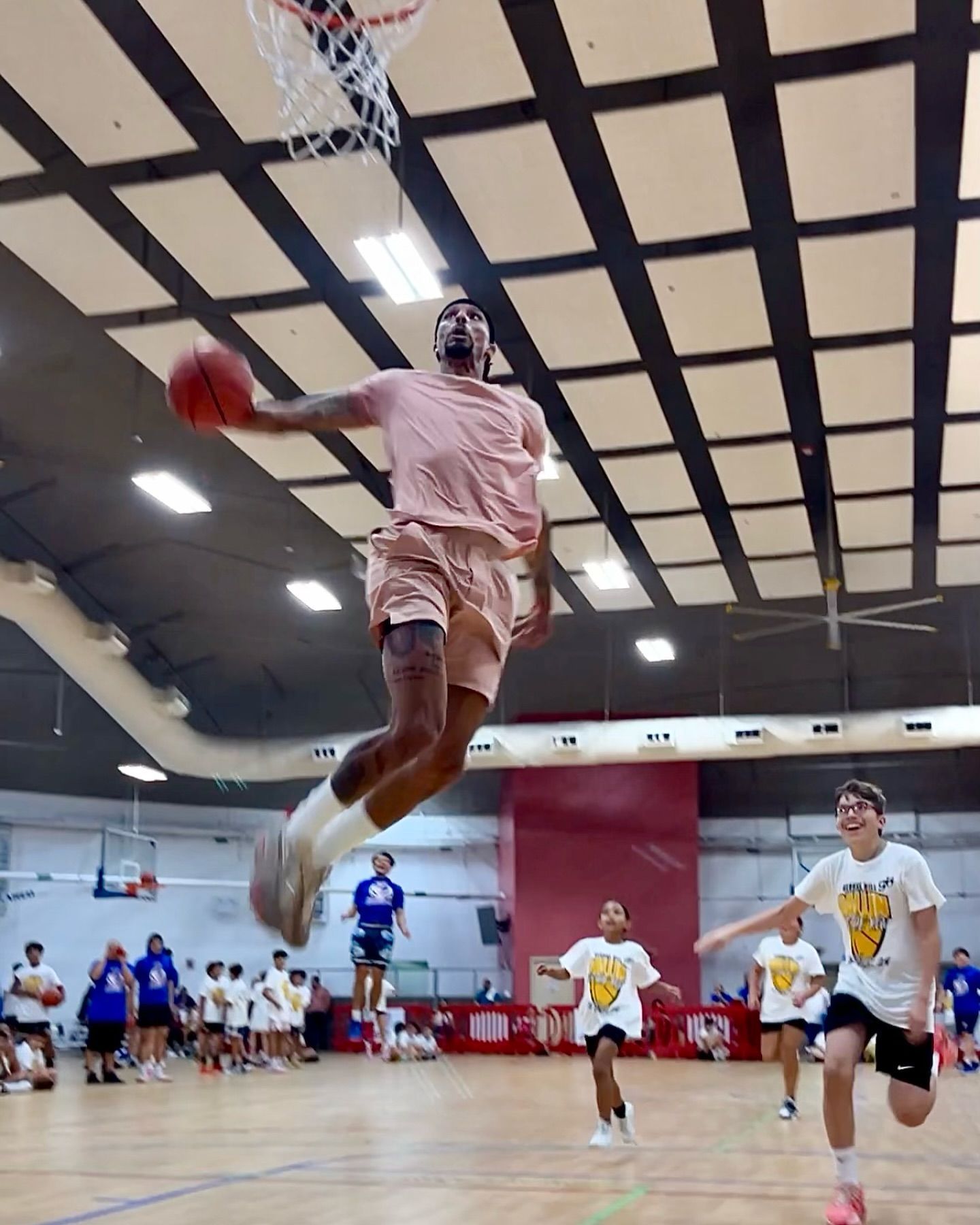 Basketball player in pink outfit dunks ball in a gym, kids watch and cheer.