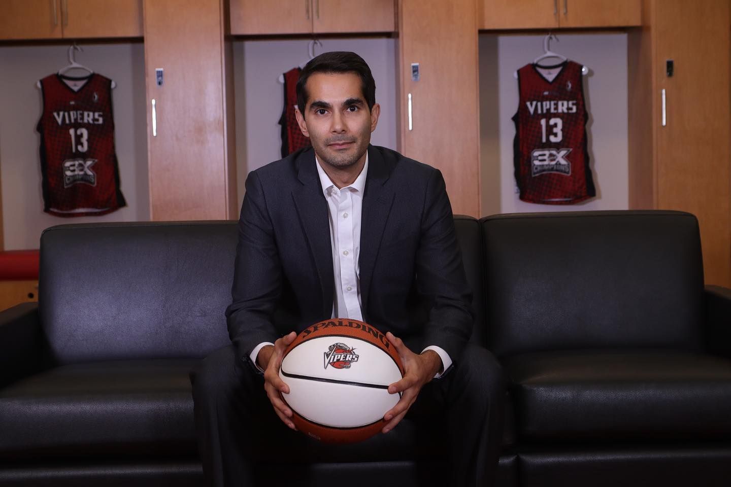 A man in a suit sits on a leather couch, holding a basketball, with two jerseys behind him.