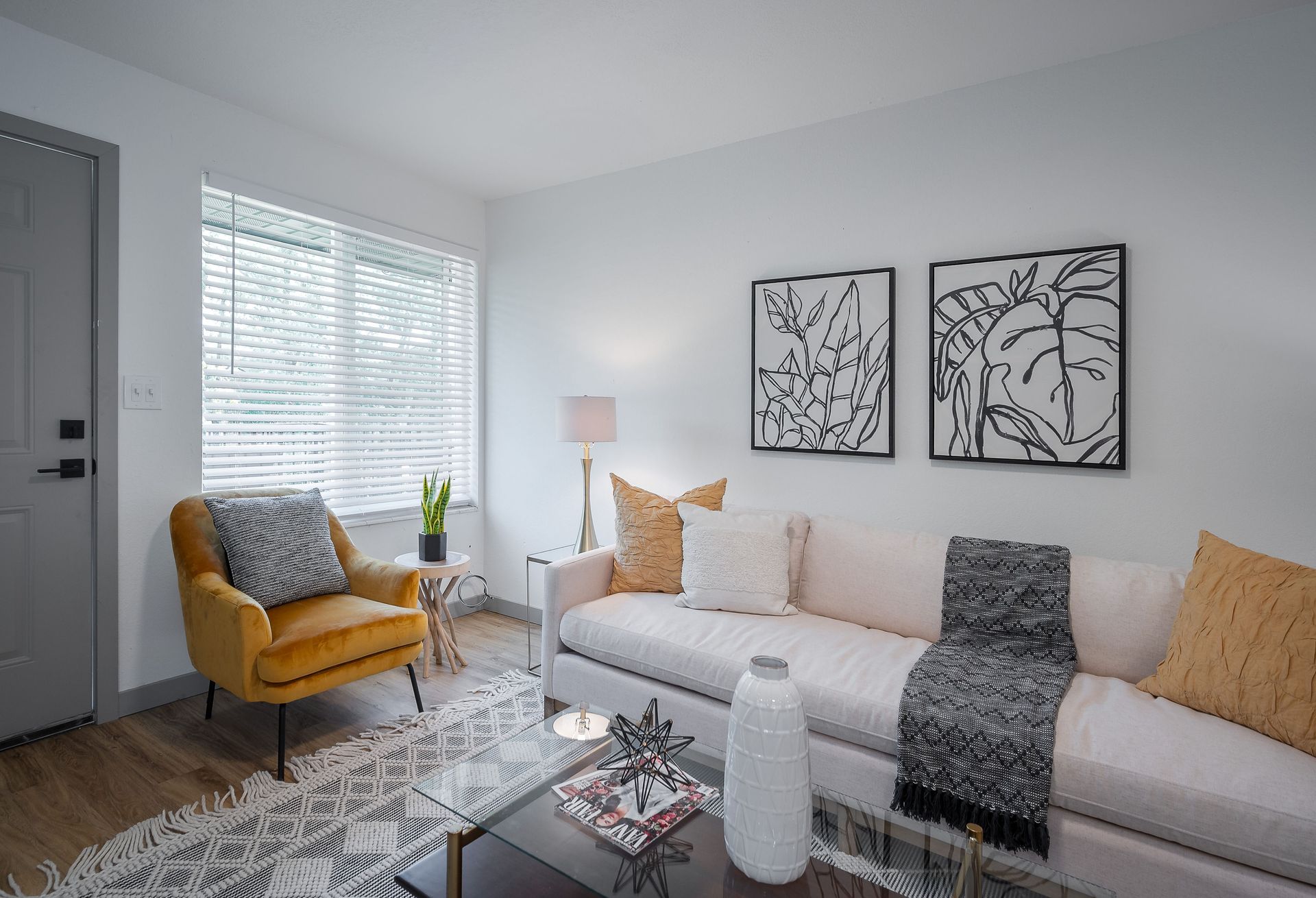 Living room with white walls, mustard chair, cream couch, abstract art, and a glass coffee table.