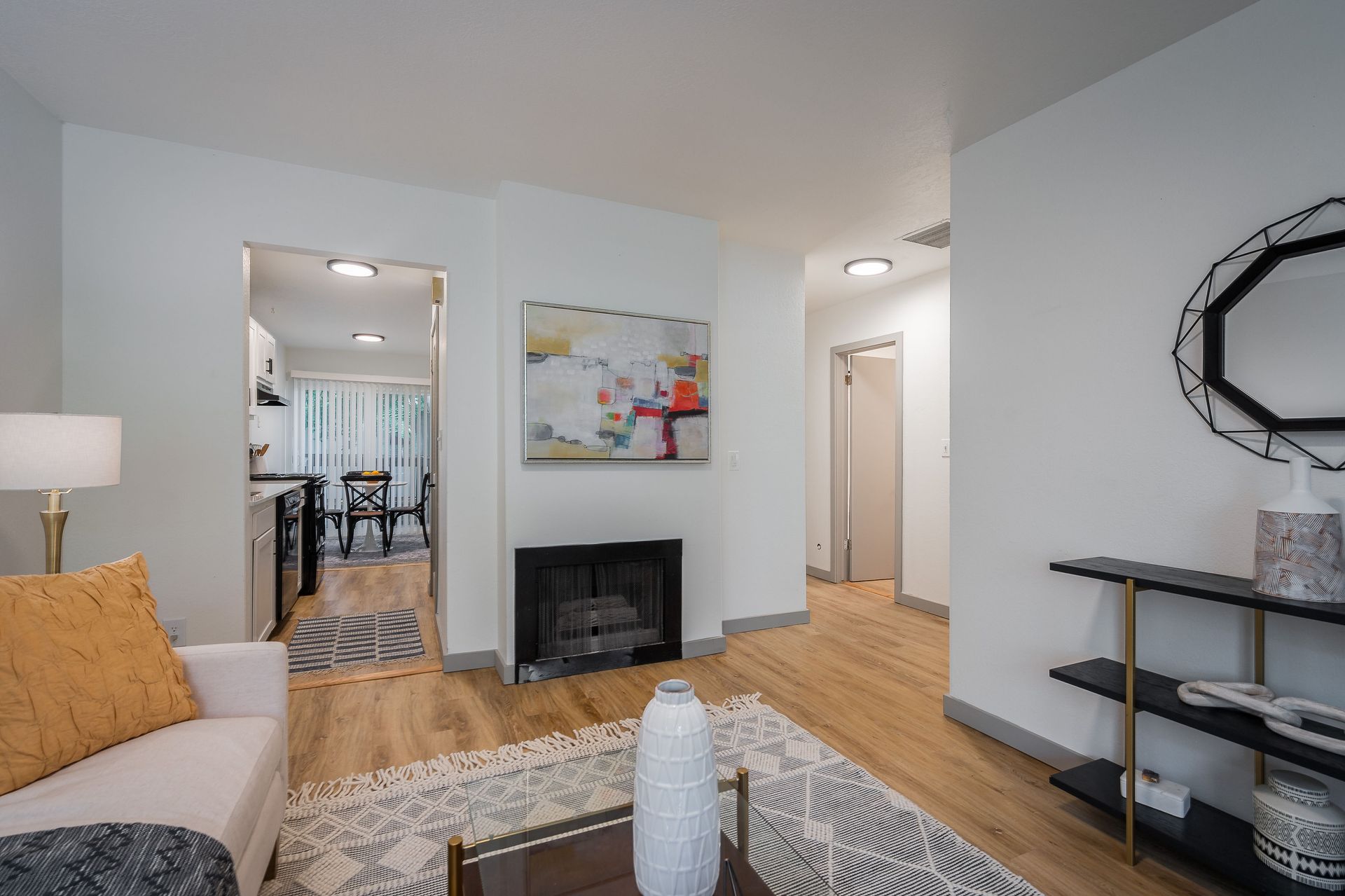 Bright living room with fireplace, leading to kitchen and hallway. White walls, wood floor, decorative furniture.