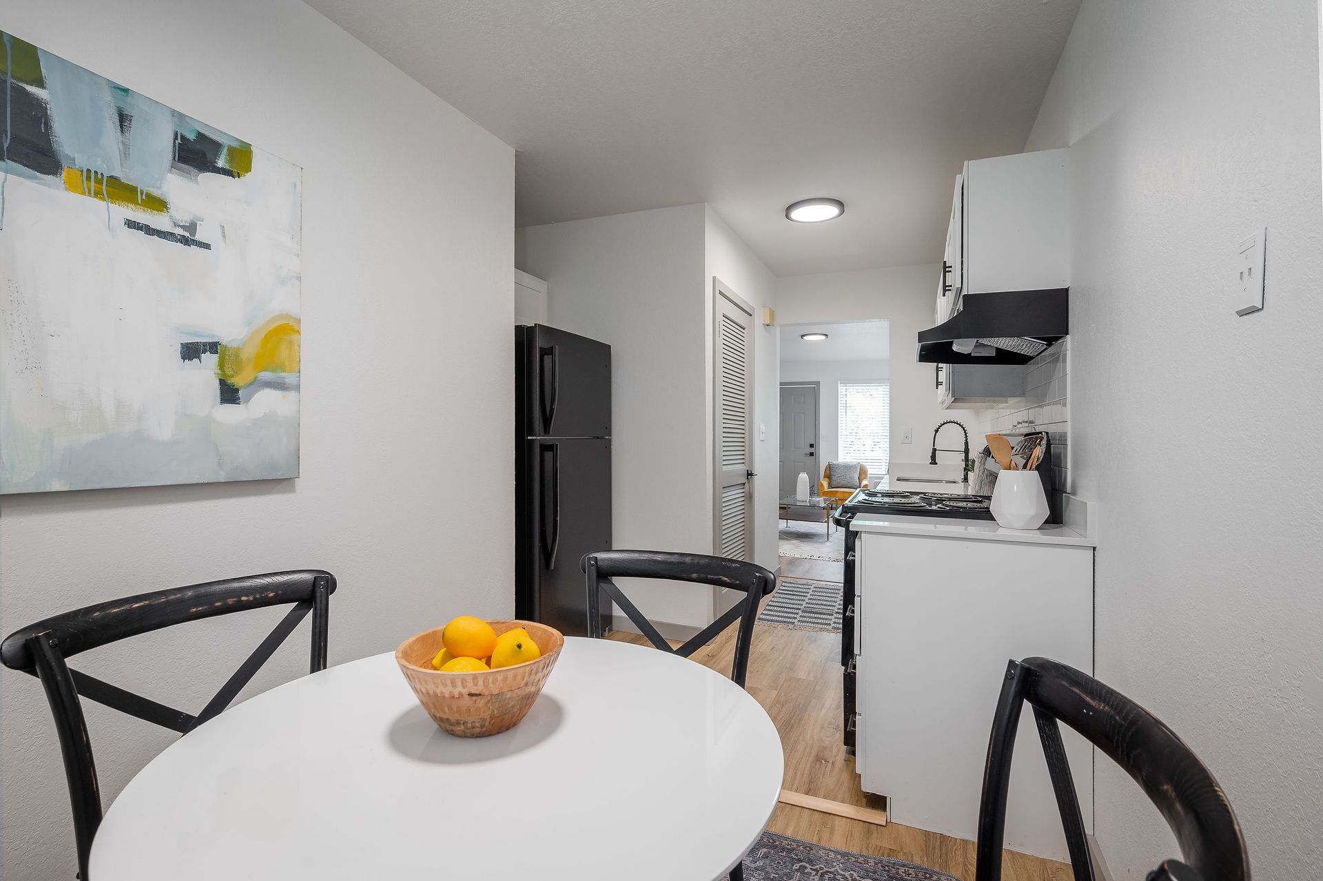 Kitchen with white table, black chairs, artwork, black refrigerator, and view into living area.