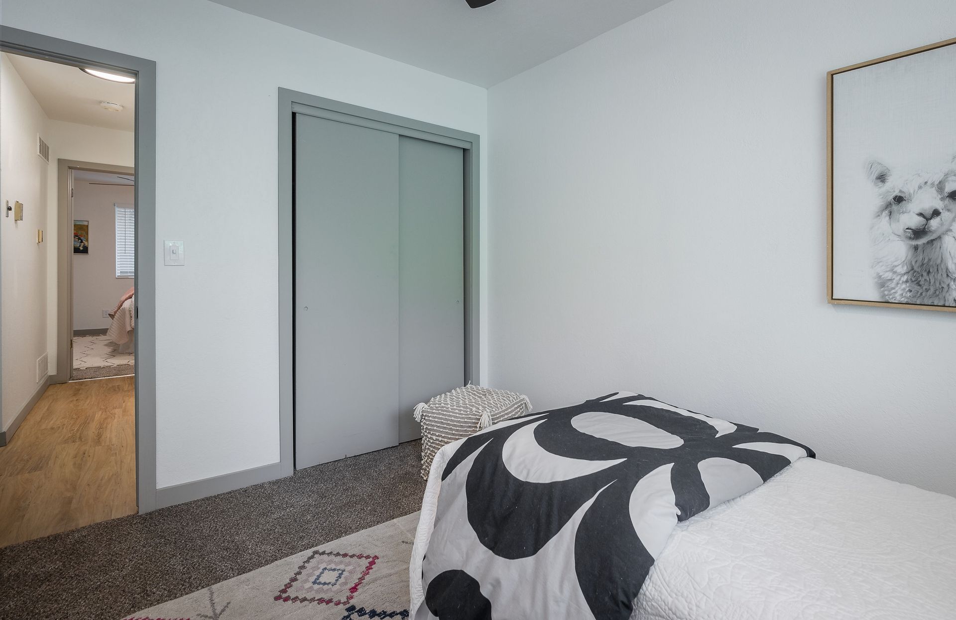 Bedroom with white walls, gray closet doors, bed with black and white patterned comforter, doorway to hallway with wood flooring.