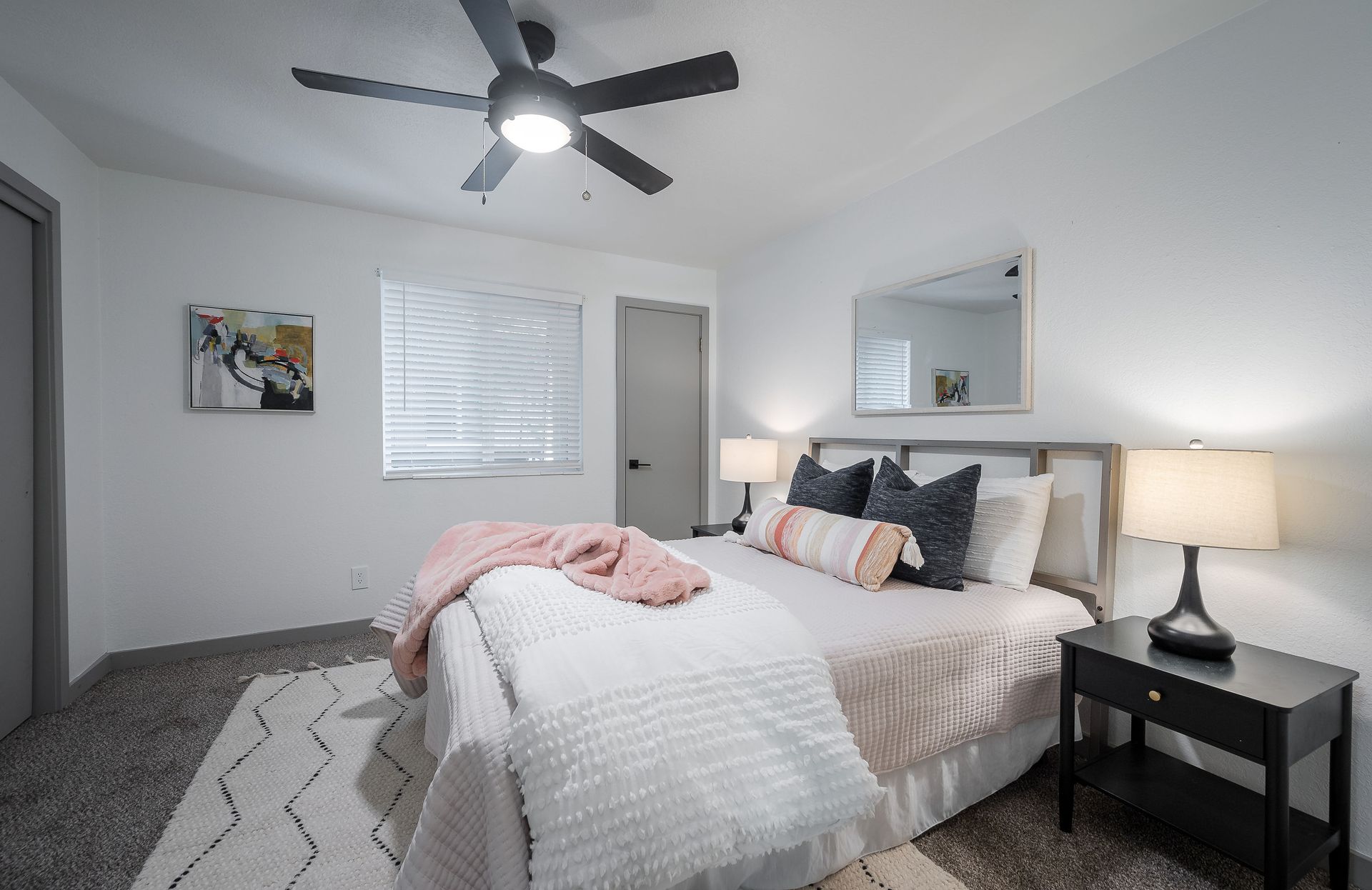 Bedroom with gray carpet, bed with pink accents, ceiling fan, and black nightstand.